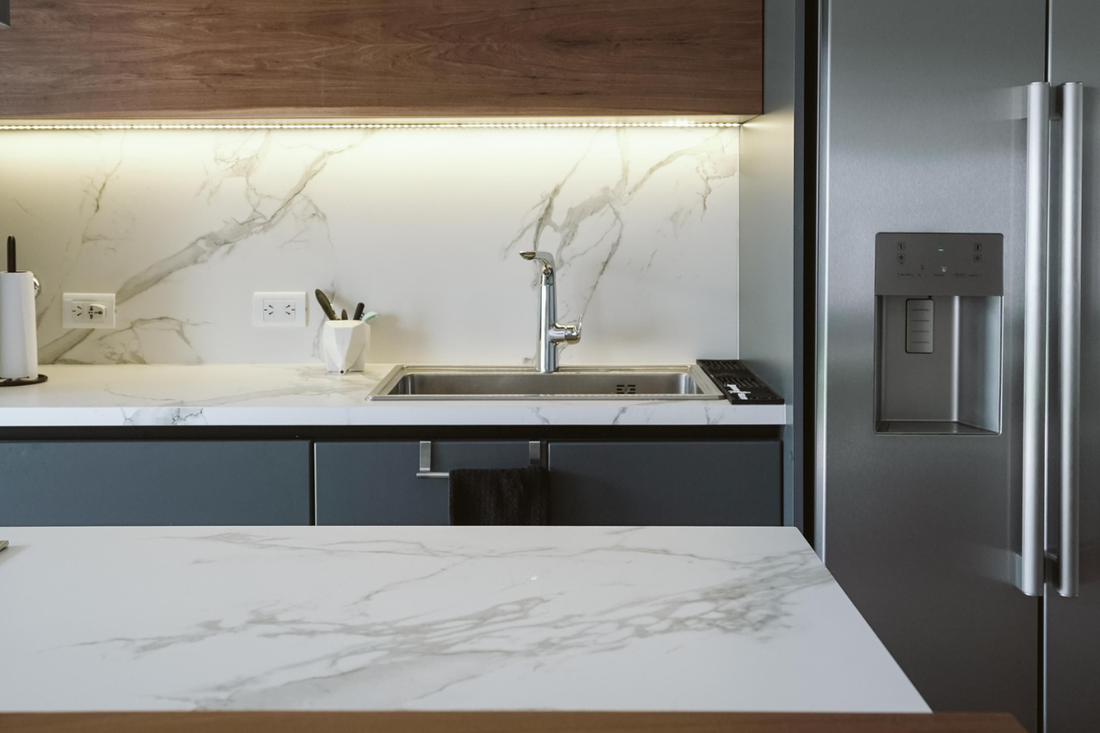 Cocina moderna con encimera y frontal de mármol blanco, muebles oscuros y frigorífico de acero inoxidable