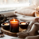Velas encendidas sobre una rodaja de madera junto a una manta beige y hojas secas, ambiente hygge