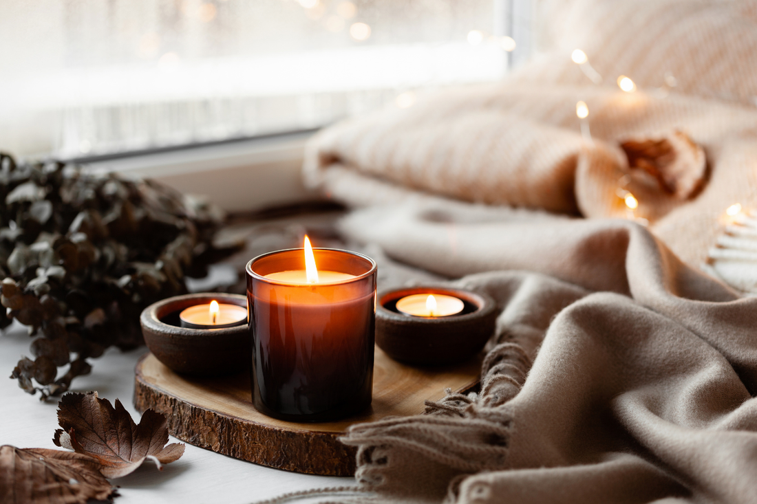 Velas encendidas sobre una rodaja de madera junto a una manta beige y hojas secas, ambiente hygge