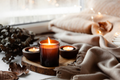 Velas encendidas sobre una rodaja de madera junto a una manta beige y hojas secas, ambiente hygge